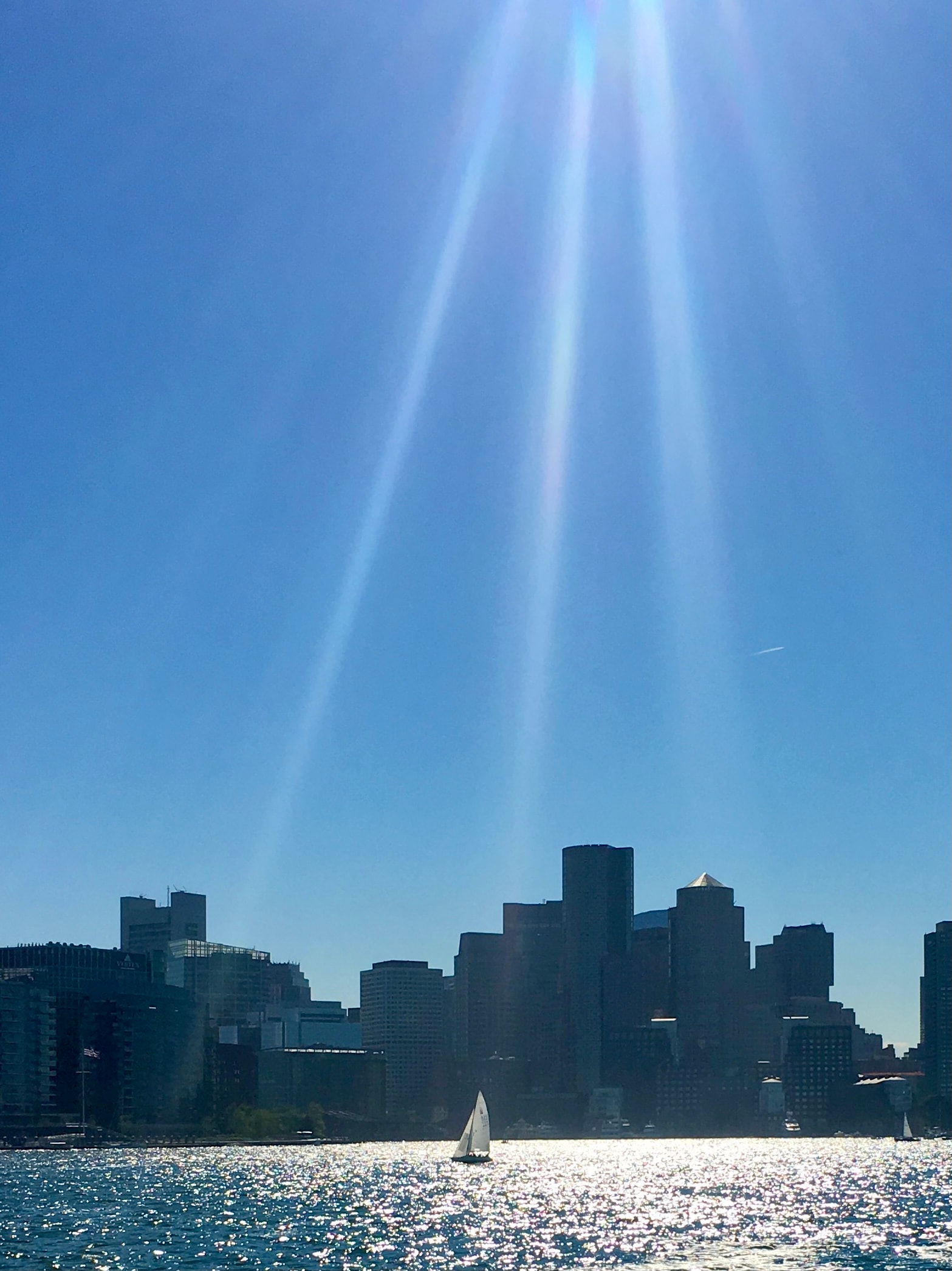 Boston city skyline from the harbor with a sailboat and sunbeams